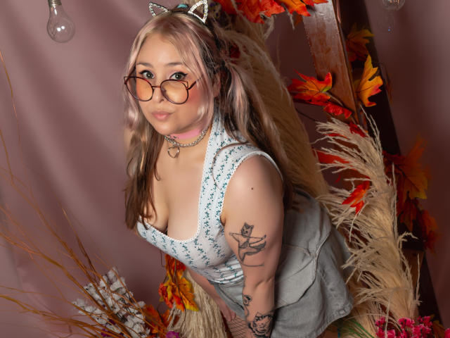 DandelionQueen with pink and blonde hair, cat ears, glasses, heart necklace, gray dress, large tits, tattoos, autumn leaves, and fluffy decor, leaning forward, smiling.