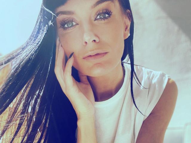 Minnieme-hot with long black hair, light skin, and green eyes, wearing a white shirt, touches her cheek with her hand, looking at the camera with a slight smile.