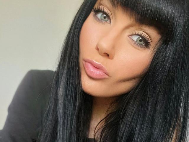 Minnieme-hot ha capelli neri lucidi, occhi verdi e labbra rosa lucide. Il suo viso è leggermente inclinato con un'espressione appena accennata. Indossa una maglia nera..