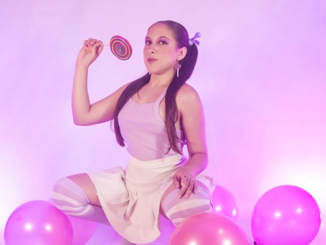 Kamie Carter, con dos largas coletas, vistiendo una camiseta rosa, una falda blanca y calcetines a rayas hasta el muslo, sosteniendo un caramelo en espiral de colores, rodeada de globos rosas..