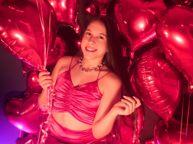 Kamie Carter, con palloncini rosa a forma di cuore scintillanti, indossa una camicetta di seta rosa e una collana di perle, sorride alla fotocamera e tiene un filo di palloncino. Ha il seno piccolo, è carina..