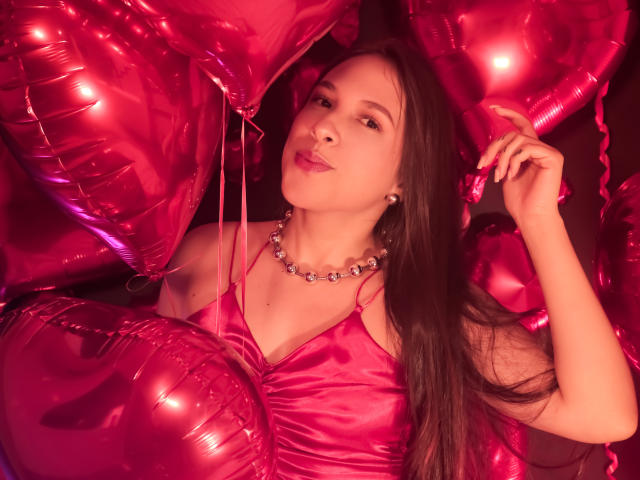 Kamie Carter, con cabello castaño largo, vestida con un vestido de satén rosa y un collar de cuentas, rodeada de globos rosas brillantes, con una expresión ligeramente afligida pero relajada y segura de sí misma..