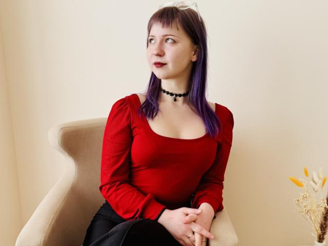 Ursula Anderson, con cabello morado, una blusa roja, un collar negro y una falda negra, sentada en una silla beige, mirando hacia un lado con una ligera sonrisa, con las manos en el regazo..