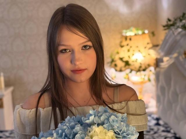 Bubochka mit langen, braunen Haaren, einem weißen Top mit Schulterpartie, die blaue und weiße Papierblumen hält, weiches Licht, dezentes Make-up, sanfter Gesichtsausdruck, warme Innenraumgestaltung..
