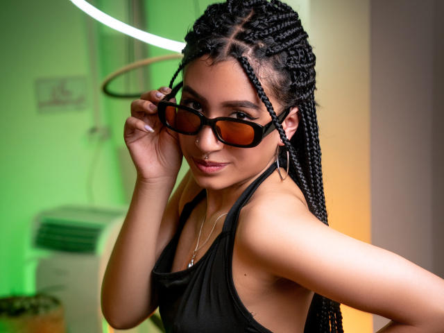 SophiaAndTony with braided black hair, wearing black glasses and a black tank top, showing her small tits, smiling, and holding her glasses with a green and yellow background.
