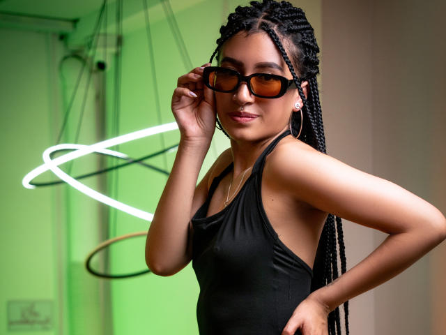 SophiaAndTony with long braids, wearing a black tank top, black sunglasses, and hoop earrings, adjusting her glasses, standing with a hand on her hip, smiling.