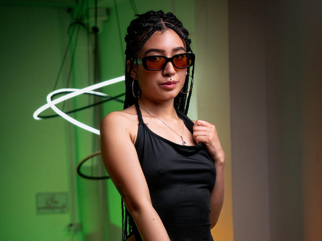 SophiaAndTony with braided black hair, brown glasses, black tank top, and hoop earrings, holding shirt with a confident smile, green and white light in background.