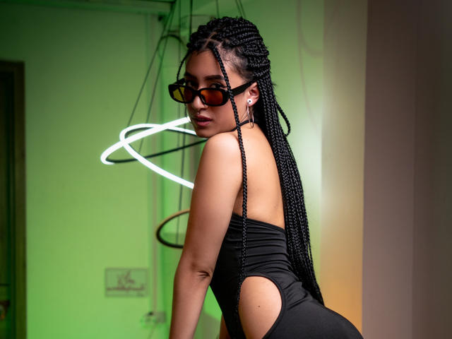 SophiaAndTony with long black braids, wearing a black cut-out one-piece, dark sunglasses, and a confident look, standing in a green-lit room with a white neon ring.