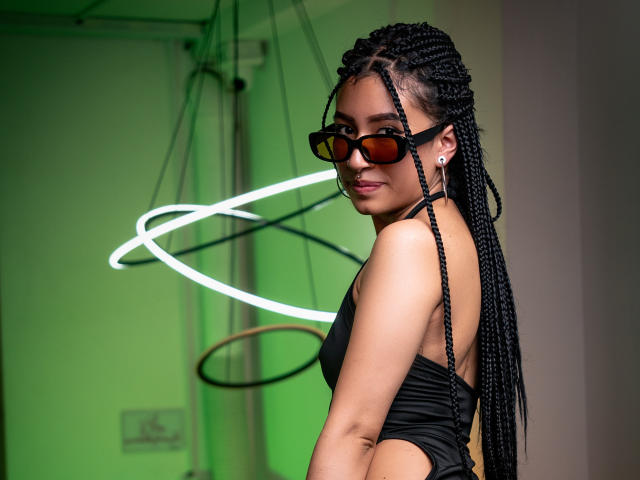SophiaAndTony with long black braids, wearing black sunglasses and a black one-piece, showing her back and smiling over her shoulder. Green light in the background.