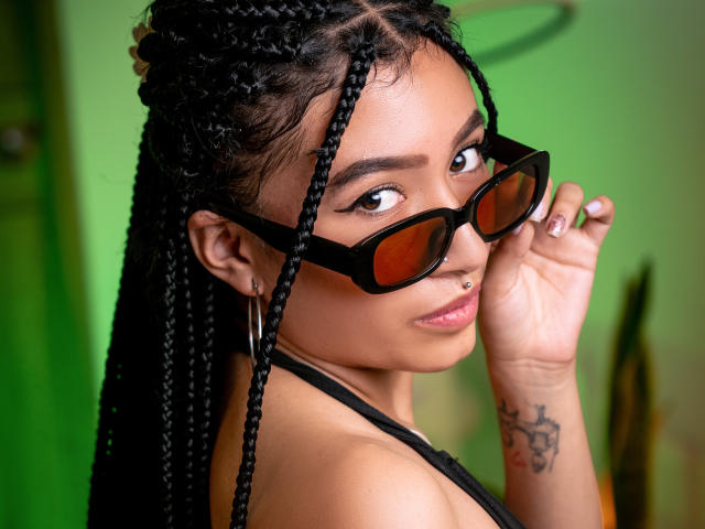 SophiaAndTony with braided black hair, brown eyes, and a nose piercing, wearing black sunglasses, showing a tattoo on her right wrist, and holding her glasses.