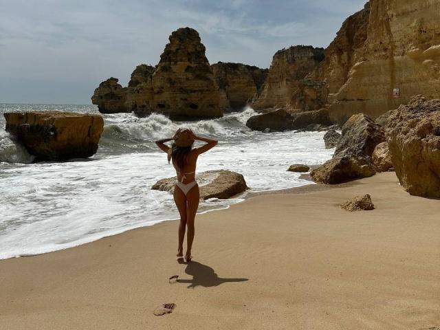 HotBabeArt with long brown hair, tan skin, and a white bikini stands on a sandy beach, hands in her hair, facing rocky cliffs and crashing waves.