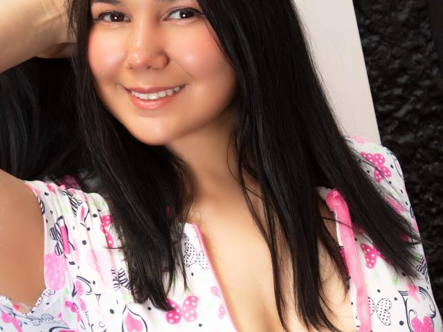 IvanaStaysi mit schwarzen Haaren, lächelnd, trägt ein blumiges Hemd in Rosa und Weiß, das ein wenig Ausschnitt zeigt, und hält sich mit einer Hand den Kopf. Ihre Brüste sind teilweise sichtbar..