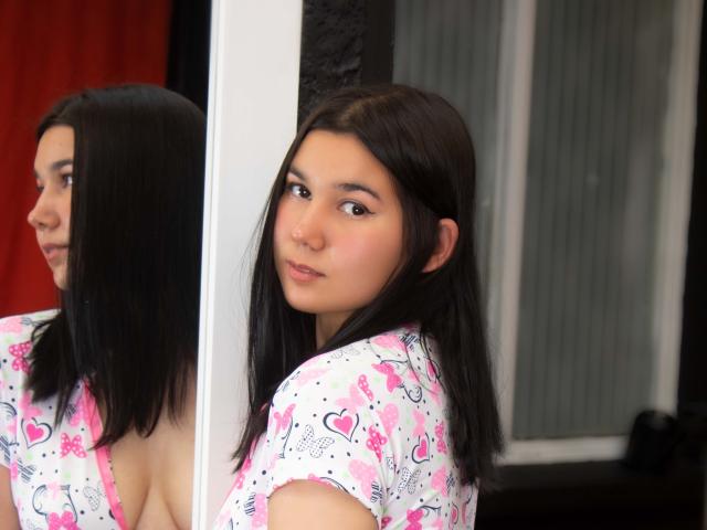 IvanaStaysi hat dunkelbraunes Haar, hellbraune Haut und kleine Brüste. Sie trägt ein rosa und weißes Oberteil mit Herzmuster und steht neben einem Spiegel, wobei sie über ihre Schulter schaut..