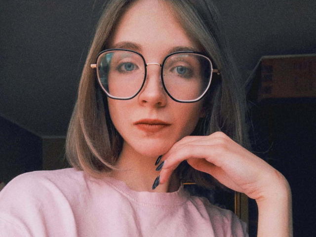 PennyDelight with glasses, pale pink sweater, shoulder-length blonde hair, and black nails, touching her chin, looking at camera with blue eyes.