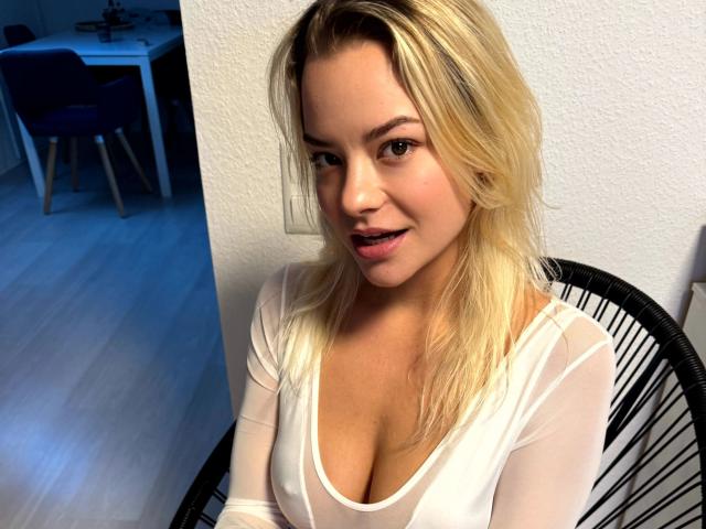 Anna-Lena-Sofia, attraktiv mit blonden Haaren, trägt ein weißes, durchsichtiges Oberteil, das ihre Brüste zeigt, lächelt und sitzt auf einem schwarzen Stuhl. Im Hintergrund ist ein blauer Schein zu sehen, der Holzboden ist sichtbar. Sie schaut in die Kamera..