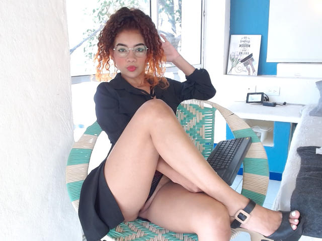 LizzaTaila with curly red-orange hair, glasses, black dress, bare thighs, black heels, and black panties, sitting on green-striped chair, hand in hair, looking at camera.
