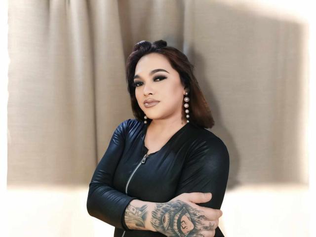 BoobsieBigCock with dark brown hair, black leather top, and pearl earrings, arms crossed, showing tattoo on right arm, looking confident. BoobsieBigCock has a sexy, attractive face.