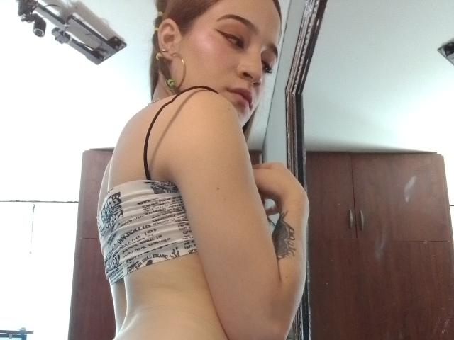 MadisonAngel with pale skin, brown hair in a bun, wearing a white and black strapless top, showing her small tits, looking over her shoulder, in a mirror selfie.