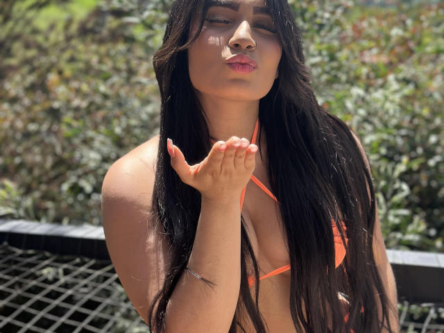 AshlyFerria, con cabello negro largo, un bikini naranja y labios rosados, haciendo pucheros con la mano cerca de la cara, la luz del sol resaltando su piel bronceada y un piercing visible en la nariz..
