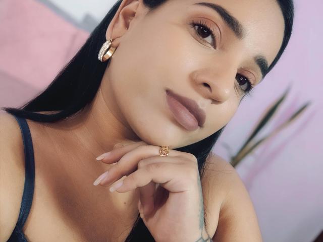 WildRachel has a cute smile, brown eyes, and long black hair. She's wearing gold earrings and a ring, with her hand near her chin. Boobs visible in black top.
