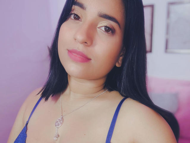 WildRachel with black hair, brown eyes, pink lipstick, blue spaghetti strap top, silver necklace with a heart charm, smiling softly, medium skin tone, cute and attractive.