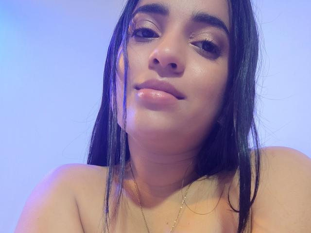 WildRachel with shiny skin, long black hair, and glossy pink lips, looking slightly to the side with a subtle smile, wearing a thin gold necklace, topless.