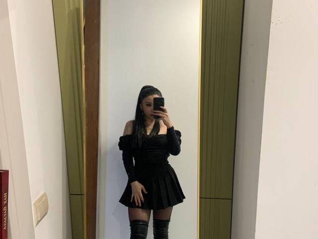 EveLovelace, con cabello negro largo, vestida con un vestido negro de corte asimétrico y botas negras hasta el muslo, se toma un selfie frente al espejo con su teléfono, mostrando su trasero desnudo..