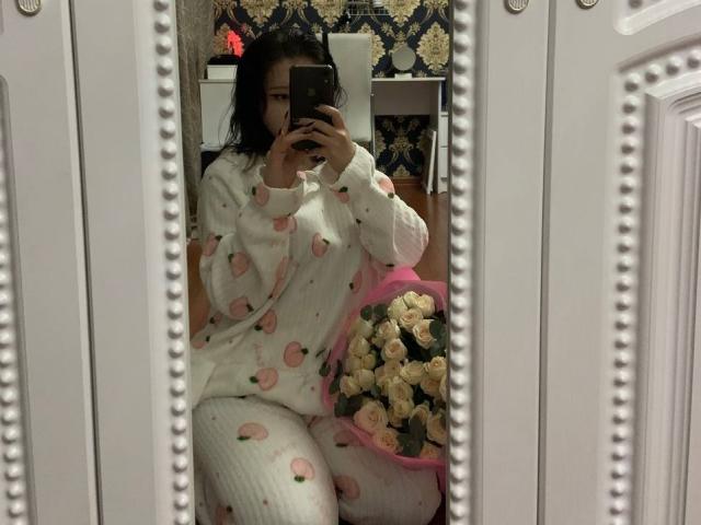 EveLovelace, avec ses cheveux noirs, vêtue d'un pyjama rose et blanc à motifs de pêches, tenant un panier rose rempli de roses blanches, prenant un selfie devant un miroir orné de perles blanches..