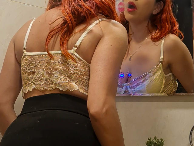 ZoeeMar met rood haar, een top van kant en zwarte broek, staat voor een spiegel en toont haar rug en de reflectie van haar borsten met een beha met blauwe steentjes. Haar lippen zijn lichtjes open..