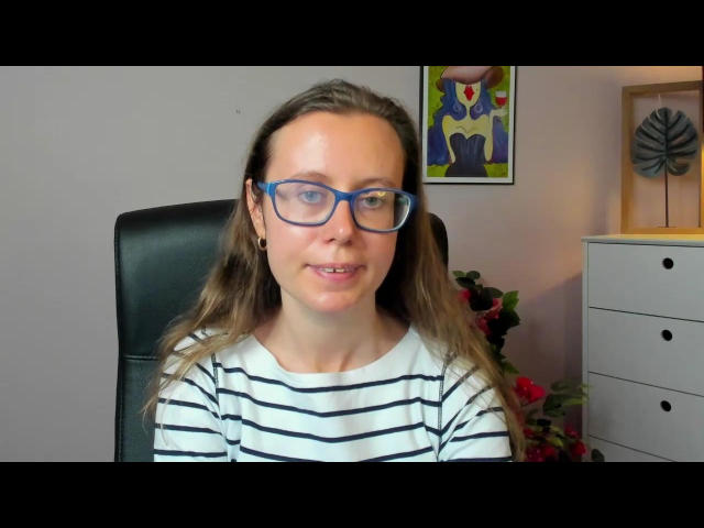 LizzySparkle met blauwe bril, bruin golvend haar, wit gestreept shirt, zittend in een zwarte stoel, lachend, lichte huid, kleine zwarte oorbellen, witte dressoir met een rode plant aan de rechterkant..