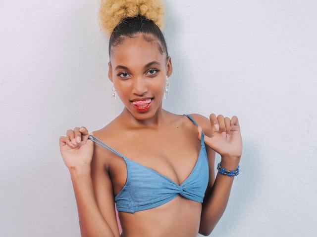 ReineElise with curly blonde hair in a bun, blue bra, small tits, smiling, holding bra straps, red lipstick, blue bracelet, white wall, looking at camera.