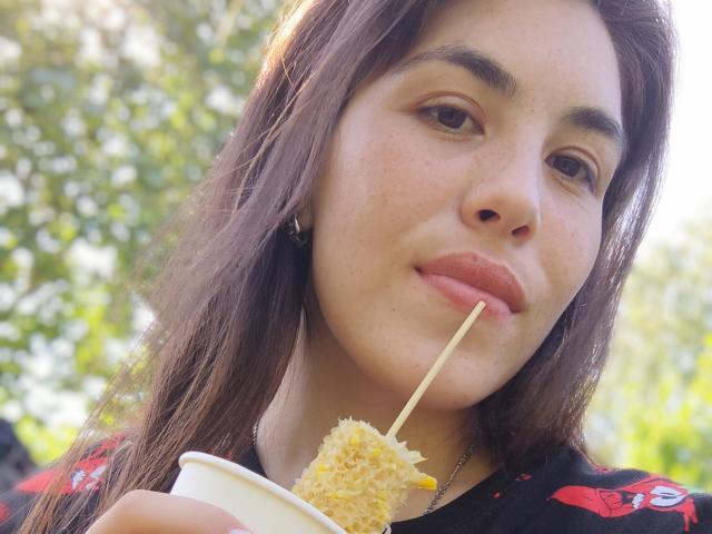 ErikaGloss with brown hair, holding a stick of fried dough, smiling with a straw in her mouth, wearing a black shirt with red patterns, in a sunny outdoor setting.