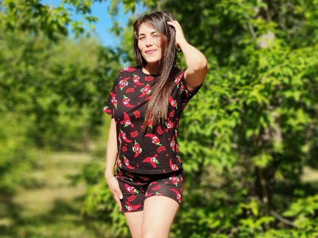 ErikaGloss with long brown hair, wearing a black red cherry patterned shirt and shorts, smiling, hand in hair, standing outdoors, green leafy background, sunny, relaxed, attractive.