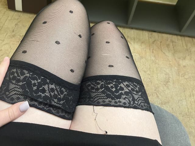 DiLaria's legs in black sheer polka dot stockings with lace tops, her pale skin and a visible small scar on her upper thigh, a hand with lavender nail polish.
