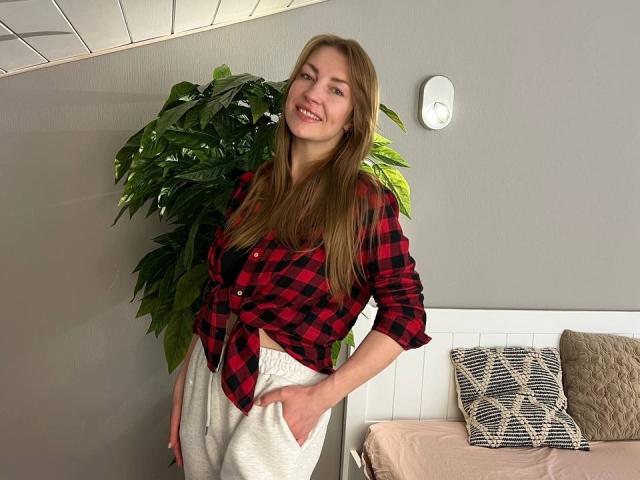 BlackMango with long blonde hair, red-black plaid shirt tied at the front, white pants, smiling, hand in pocket, standing next to a green plant and a bed with pillows.