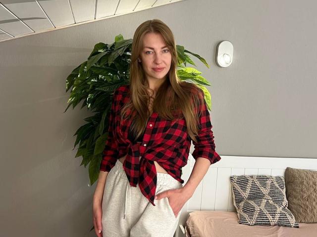 BlackMango has long brown hair, wears a red black checkered shirt tied at the front, and white high-waisted pants, standing with hand in pocket, smiling softly.