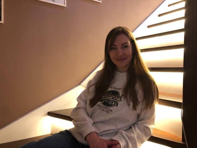 BlackMango with long brown hair, smiling, wearing a gray "Kalev" sweatshirt and blue jeans, sitting on a lit staircase, hands resting on her knee.