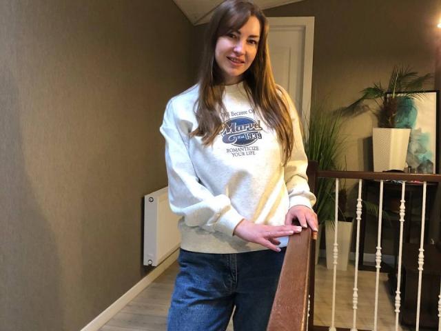 BlackMango, with long brown hair, wears a gray "Pulse" sweatshirt and blue jeans, standing by a wooden railing, smiling, hands resting on the rail.