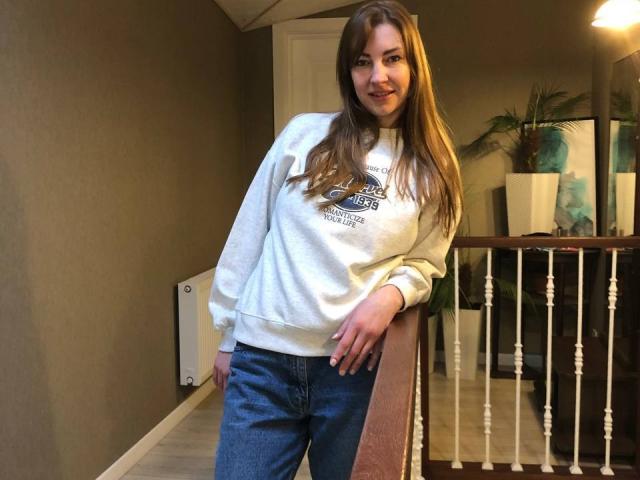 BlackMango with long brown hair, white sweater, and blue jeans, leaning on a wooden railing, smiling, hands resting on her hip, standing in a dimly lit hallway.