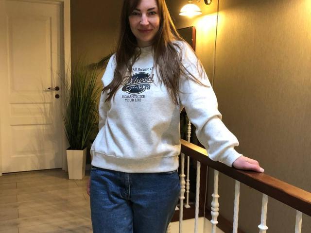 BlackMango with long brown hair, wearing a white sweatshirt and blue jeans, standing by a wooden railing, smiling, hand on railing, indoors, under warm light.