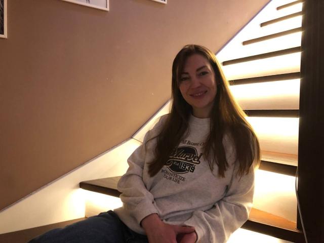 BlackMango, sitting on a lit staircase, wearing a grey sweatshirt and blue jeans, smiling with long brown hair, her ass slightly visible on the step.