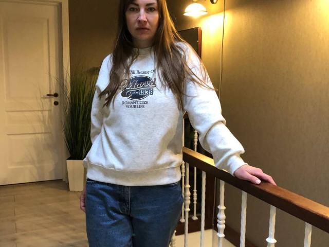 BlackMango stands with a neutral expression, wearing a white "Champs" sweatshirt and blue jeans, her hand on a wooden railing, brown hair, and a slight frown.
