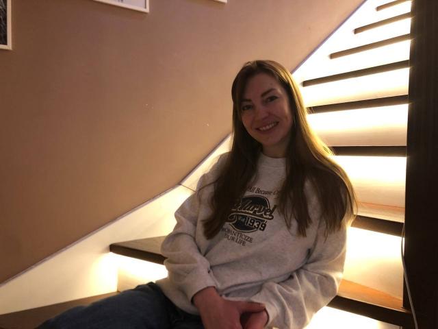 BlackMango, a cute woman with long brown hair, is sitting on a lit staircase. She's wearing a gray "Raiders 1984" sweatshirt and blue jeans, smiling.