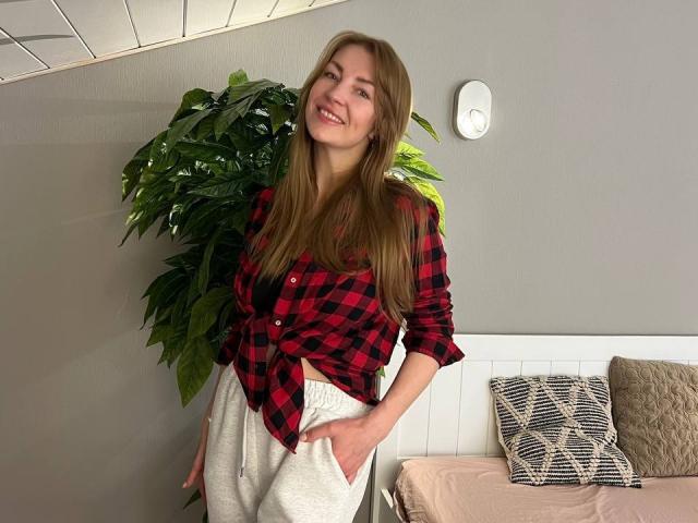 BlackMango with long brown hair, red plaid shirt tied at the front, and white sweatpants, standing with hands in pockets, smiling. Potted plant and bed with pillows behind.