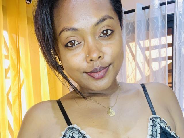 Cathy69s with dark skin, brown eyes, and a small gold necklace, smiling at the camera. She's wearing a black and white lace top, showing her small tits.