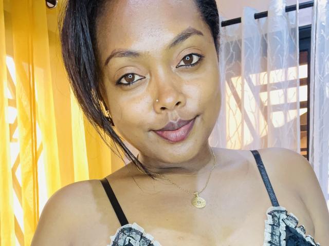 Cathy69s has brown eyes, medium brown skin, and a slight smile. She's wearing a black and white patterned top with thin straps. She has a gold necklace.