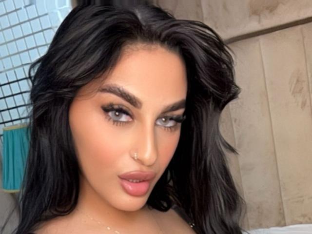 SophyAngels tiene el cabello oscuro y ondulado, un piercing en la nariz y labios rosados y carnosos, con pestañas largas y gruesas, mirando directamente a la cámara con una expresión sensual..
