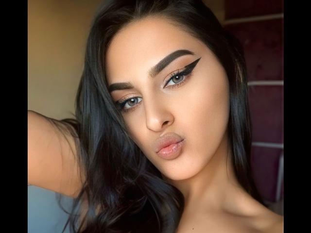 SophyAngels tiene el cabello oscuro y ondulado, cejas negras y gruesas, y un delineado de ojos alado y definido. Sus labios carnosos están ligeramente entreabiertos, mostrando un tono rosado y brillante. Tiene un rostro lindo y atractivo..