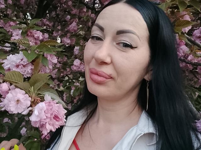 MirandaHott69 mit schwarzen Haaren, pinkem Lippenstift und Eyeliner im "Winged"-Stil, die die Lippen zu einem Schmollmund formt und die Zunge leicht herausstreckt, hält rosa Kirschblüten, trägt eine weiße Jacke und goldene Ohrringe..