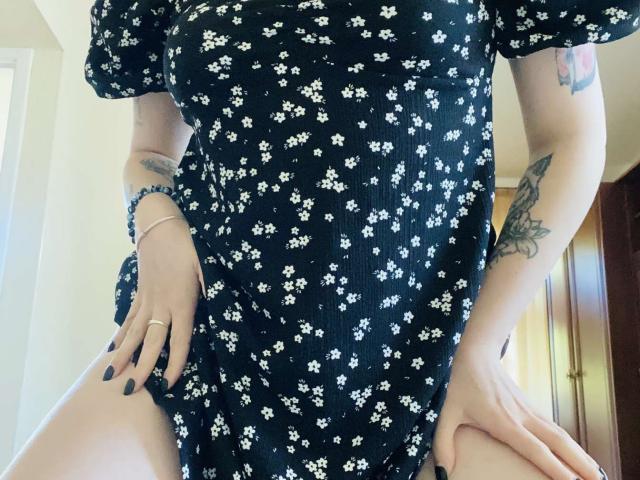 KarKutie wearing a black floral dress, showing her thighs, hands on her hips, tattoos on her arm, black nail polish, bracelet on her wrist, in a well-lit room.